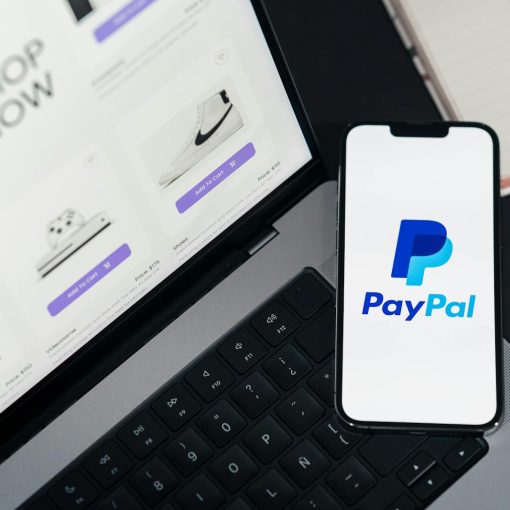 Smartphone displaying PayPal logo next to laptop with online shopping site open.